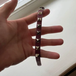 Valentino Armband - Snyggt armband i mörkröd färg med silverfärgade nitar. Perfekt accessoar för att ge din outfit en edgy touch. Armbandet är smalt och justerbart för att passa de flesta handleder.