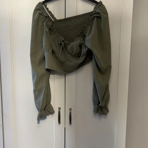 Olivgrön offshoulder topp - Snygg olivgrön offshoulder topp med långa ärmar och smockdetaljer. Toppen har en knytning framtill och volanger vid ärmsluten för en extra touch. Perfekt för en trendig look.