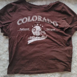 Brun t-shirt med tryck - Säljer en brun babytee med tryck av texten 'Colorado National League' och en hjälm. 