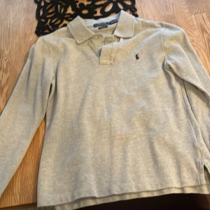 Grå långärmad pikétröja från Ralph Lauren - Säljer en stilren grå långärmad pikétröja från Ralph Lauren med klassisk krage och knappdetalj. Perfekt för en avslappnad men ändå snygg look. 🐎Storlek 14-16 år