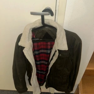 Olivgrön jacka med teddyfoder från Be Edgy - Säljer en olivgrön jacka från Bleed med mysigt vitt teddyfoder. Jackan har en rutig insida i rött och blått samt dragkedja framtill. Perfekt för kyliga dagar! Knappt använd 