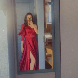 Bröllop klänning  Röd långklänning med slits - Elegant röd långklänning i satin med en axel och en dramatisk slits. Klänningen har ett bälte i midjan som framhäver figuren och ger en stilren look.