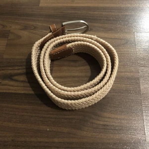 Beige flätat bälte - Snyggt beige flätat bälte med silverfärgat spänne och bruna läderdetaljer. Perfekt för att ge en stilren touch till din outfit.