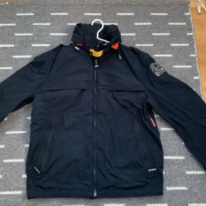 Mörkblå  jacka från Parajumpers - Säljer en svart jacka från Parajumpers med orange detaljer och dragkedja. Jackan har en broderad logga på ärmen och är långärmad. Perfekt för höst och vår. Jackan är tunn och OBSERVERA att det finns ett litet hål 🕳️ på vänster underarm