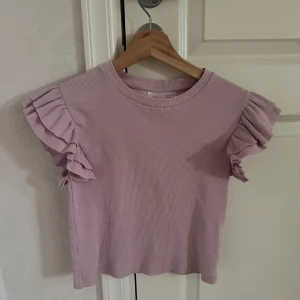 Ljusrosa ribbad topp med volangärmar  - Söt ljuslila ribbad topp med volangdetaljer på ärmarna. Är från Gina Tricot och i storlek 146/152, bra skick. Priset kan diskuteras. 
