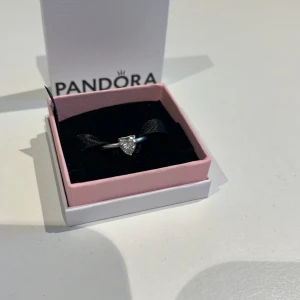 Silverring med hjärtformad sten från Pandora - Elegant silverring från Pandora med en gnistrande hjärtformad sten i mitten. Perfekt för att ge en touch av glamour till vilken outfit som helst. Levereras i en fin presentask. Använd ett tag därav det låga priset, är ändå i fint skick. 