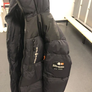 Svart dunjacka från Sail Racing - Snygg svart dunjackafrån Sail Racing med coola detaljer och logotyper. Perfekt för kyliga dagar med dragkedja och praktiska fickor. Passar bra för både höst och vinter.