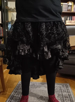 Svart kjol med volanger  - Svart kjol med glansig underkjol och genomskinlig, mönstrad överkjol. Dragkedja i midjan. Kort fram, ca 35cm, halvlång bak ca 70cm. Knappt använd.