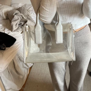 Genomskinlig väska - Väska från zara med grått ormskinsliknande mönster på handtag och botten. Resten är genomskinligt. Ok skick, blå fläck som inte går bort, samt en lös söm på handtaget. Tillhörande axelrem