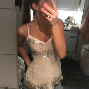 En beige tröja med paljetter på - En vit/beige topp som sitter tajt runt bröstkorgen och e v-ringad. Har paljetter på sig. Min mammas från 2000s