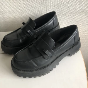 Svarta loafers storlek 39 - Svarta loafers i skinn immitation. Tidigare satt det en kedja på ovansidan som är borttagen. Sulan är lite sönder på höger innersida. Sköna och mjuka. Perfekta att gå ut i!🖤
