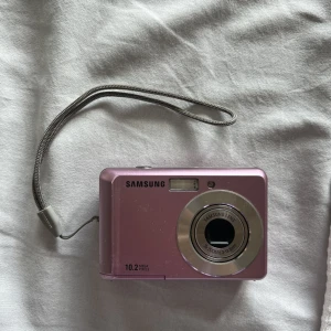 Digitalkamera  - Rosa kompakt digitalkamera Samsung. Trasig på locket där batterier & minneskort ska in men går att sätta ihop med tejp. Fungerar förövrigt utmärkt. Kommer ej mer till användning. Minneskort + AA batterier ingår ej