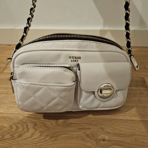 Vit axelväska Guess - Hej! Säljer denna, den är använd, vilket man kan se på baksidan av väskan. Det är missfärgning av jeans, har ej testat att tvätta bort det. Väskan är från Guess och har både inne samt utefack!