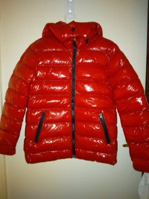 HELT NY & OANVÄND snygg röd glansig vinterjacka, dam. Strlk: Large. - HELT NY & OANVÄND snygg röd glansig vinterjacka, dam.  Strlk: Large, men passar även någon som har Medium då ganska liten Large. Är ej dun men vadderad/täckjacka som verkar ganska värmande. Har fickor med dragkedjor samt avtagbar luva. Slutsåld i butik.