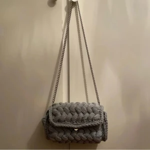 Väska - Grå virkad flätväska gjord på bomullsgarn från textilindustrins rester. I denna får du plats med det absolut nödvändigaste (mobil, nycklar och plånbok). Att ha i åtanke är att den väger kring 1500g! Mått: längd:27,5cm ,höjd:17cm ,djup:6,5cm 