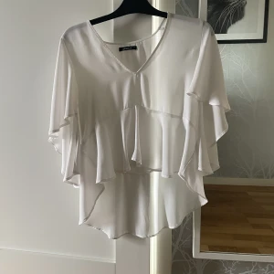 Vit blus - Vit blus som ser lite oversized ut men ändå anpassad för en storlek S. Ärmarna är väldigt vida (se bild 2) och runt midjan ett typ av ”svall” av tyg som formar sig vackert runt kroppens former.