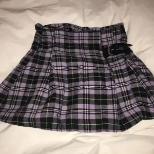 En kort kjol som är lila och svart med en liten ”bälte” detalj. Som ny. Sitter fint. Märke okänt tyvärr