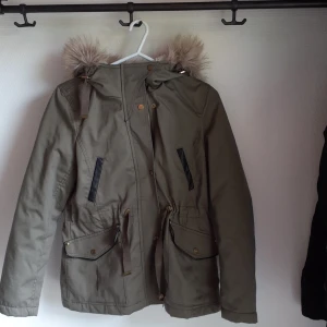 Kakigrön höstjacka med päls och läderdetaljer! - Kakigrön höstjacka med fodrad innerluva och fuskpäls runt luvan. Har svarta läderdetaljer, dragsko i midjan.