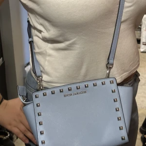 Michael Kors väska. - En äkta baby blå Michael Kors väska med nitar, endast använd ett fåtal gånger och är i väldigt bra skick. Perfekt som första märkesväskan! Med väskan tillkommer dustbag. Bandet är justerbart och inne i väskan finns en tunn ficka och ett nyckelband.