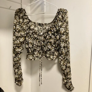 Croppad blommig singoallatopp - En oanvänd och väldigt fin croppad singoallatopp med dragsko vid bysten. Resår och volang nedtill. Svart med blommigt mönster i vitt och mörkare beige. Prislappen är kvar. Strl 42 men något liten i storleken. Köpt för 299kr.