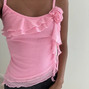 Rosa linne / top - Jättesött Rosa snedskuret / asymmetriskt linne med blomma och volang / volanger . Justerbara axelband.  Storlek Small  Aldrig använd, helt ny! 🌸💕