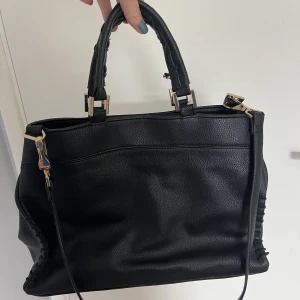 Handväska - Svart handväska från toshop som är i väldigt fint skick. Höjd 27 cm, bredd 37 cm. Coola detaljer på handtagen och guld detaljer. Finns flera fickor och avtagbart långt band som ger dig möjlighet att ha väskan ”cross body”. Pris kan diskuteras.