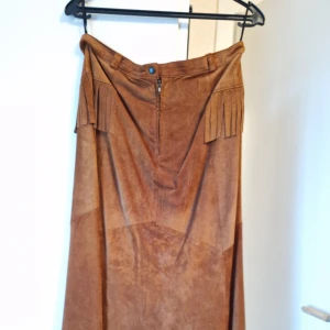 Moccakjol - Fin klockad kjol i äkta mocca. Längd 90 cm. Midjemått 80 cm. Storlek 42. I skick som ny, knappt använd. Med fransar. 