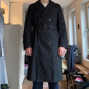 Svart trenchcoat herr, storlek 40R - En svart trenchcoat utan märke. Storlek 40R (Regular passform). Insidan har ett tillägg som gör trenchcoaten varmare och som tas av och på med en dragkedja. Använd i gott skick, inga defekter.