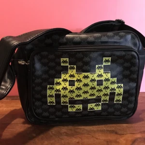 Space invaders axelväska  - Helt ny väska med retro spelet Space Invaders motiv.   Svart väska med gult/lime motiv i storlek 30*24*10cm.   Märket på väskan är SkyLine och ord.pris i butik ca 400:- Axelremsväska som kan hämtas upp i Enskededalen eller skickas mot fraktkostnad.  Gaming Retro tv-spel dator väska