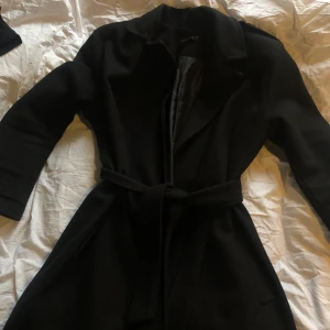 Svart Varm Kappa - Köpt från boohoo.com, använd endast tre-fyra gånger och är som ny. Fickor på sidorna, skärp som knyts vid magen och knappspänne vid armarna och axlarna