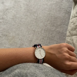 DANIEL WELLINGTON KLOCKA  - Säljer min oanvända Daniel Wellingtonklocka med uret i roseguld och bandet i tyg av färgerna vitt, rosa och marinblått.