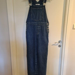 Hängselbyxor - Oanvända hängselbyxor. Denim material, fyrkantig halsringning, justerbara axelremmar, vida ben, en bröstficka, sidfickor, bakfickor, knappstäninging på sidorna. Ordinare pris 399kr