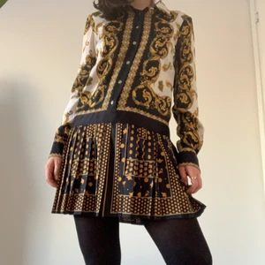 Mini shirt dress with pattern - Shirt dress with pattern from Zara. The top part of the dress is a shirt design, with buttons all up the torso and buttoned relaxed sleeves. The bottom is a pleated skirt that reaches below mid-thigh. The material is a polyester blend but feels very soft and silk-like. This dress is an excellent party dress and goes great with a pair of thigh-high boots. The collar is accessorized with a nice gold detail button, which matches the buttons on the sleeves. In good condition, all buttons attached.