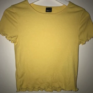 Ribbad T-shirt strl. S - En gul ribbad, lite kortare t-shirt i storlek S. Detaljer på ärmslut och längst ner på tröjan. 