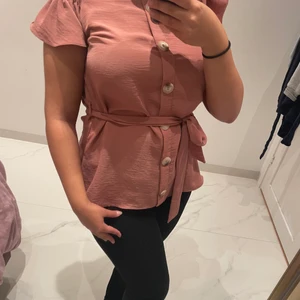 Blus - Off shoulder rosa blus med en liten volang vid båda axlarna, knappar och ett knyt band vid midjan.