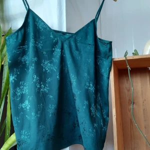 Grönt satin linne - Ett grönt linne med blommor i satinmaterial. Fint att ha sol det är eller över något annat. Skulle säga att den passar XS - M typ. Är mer emeraldgrön i verkligheten än vad som syns på bilderna