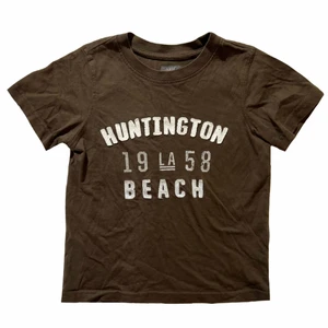 Brun babytee - Söt brun babytee med text på framsidan. Finns ett litet hål på baksidan, annars är den i fint skick. Barnstorlek, men skulle säga att den är som en XXS.