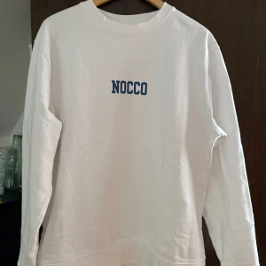 Vit sweatshirt från NOCCO - Säljer en vit sweatshirt från NOCCO med blå logga tryckt både fram och bak. Tröjan har rund halsringning och långa ärmar. Perfekt för en avslappnad dag eller till gymmet. 