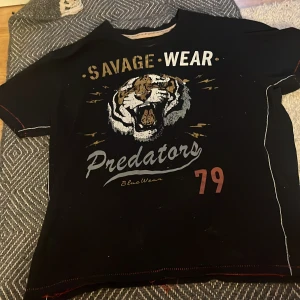 Svart t-shirt med tigertryck från BlueWear - Svart t-shirt från BlueWear med stort tigertryck och texten 'Savage Wear Predators 79' på framsidan. T-shirten har korta ärmar och normal passform. Perfekt för dig som gillar djurmotiv och streetstyle. Andvänt 1 gång:)) 