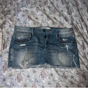 Jeanskjol med nitar från Abercrombie & Fitch - Säljer en blå jeanskjol från Abercrombie & Fitch med slitna detaljer och rå kant nertill. Kjolen har nitar vid fickorna både fram och bak samt dekorativa sömmar på bakfickorna. Perfekt för en cool och avslappnad stil.