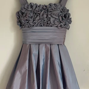 Lila cocktailklänning med volangdetaljer - Jättefin lila klänning med breda axelband och massor av blomliknande volanger upptill. Klänningen har markerad midja som kan justeras med banden i ryggen, rosett i ryggen och lite puffig nertill. Passar perfekt för bal eller fest! 💫