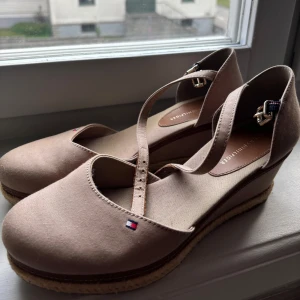 Beige sandaler med kilklack från Tommy Hilfiger - Snygga beige sandaler från Tommy Hilfiger med kilklack och justerbart spänne runt vristen. Skorna har rund tå och en liten logodetalj framtill. Perfekta för vår och sommar! Dessa är använda väldigt få gånger och har imprensip inget tecken på användning ( enbart en liten vit fläck som jag tror är möjligt att få bort.)