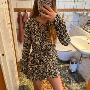 Leopardmönstrad klänning från Motel - Säljer en kort leopardmönstrad klänning från Motel med långa ärmar och volangdetaljer. Klänningen har knytning vid halsen och en smickrande passform med markerad midja. 