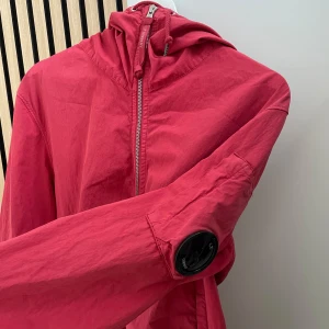 Röd vindjacka från C.P. Company - Säljer en röd vindjacka från C.P. Company med huva och dragkedja framtill. Jackan har en ikonisk linsdetalj på ärmen och två fickor med dragkedja. Str M