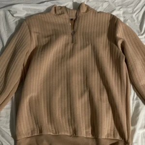 Beige ribbad tröja med dragkedja - Säljer en beige ribbad tröja med halv dragkedja framtill och ståkrage. Tröjan har långa ärmar och är tillverkad i ett mjukt material som passar perfekt för kyligare dagar och går också bra för varmare dagar