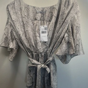 Grå ormönstrad omlottblus med knytskärp - Snygg omlottklänning i grått och svart ormmönster. Klänningen har korta, vida ärmar och ett knytskärp i midjan som ger en fin siluett. Perfekt för dig som gillar trendiga mönster och vill sticka ut.