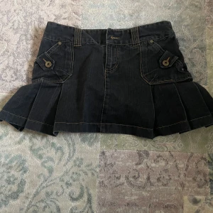 Svart jeanskjol med fickdetaljer - Cool svart jeanskjol med markerade sömmar och två fickor framtill med lock och knappdetaljer. Kjolen har veckade partier på sidorna för extra volym och bälteshällor i midjan. Perfekt för en avslappnad och trendig look.  Tyvärr för liten!