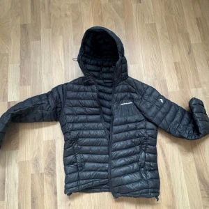 Peak Performance dunjacka med huva – stl L (svart) - Lätt och varm dunjacka från Peak Performance i svart. Perfekt som mellanlager eller ytterjacka året runt, och smidig att packa med vid friluftsliv eller resa.