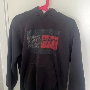 Svart hoodie från Tommy Hilfiger - Säljer en svart hoodie från Tommy Hilfiger med stor logga framtill i grått, rött och vitt. Tröjan har huva och magficka, samt långa ärmar med ribbade muddar. Perfekt för en avslappnad stil. Trycket är urtvättat och slitet