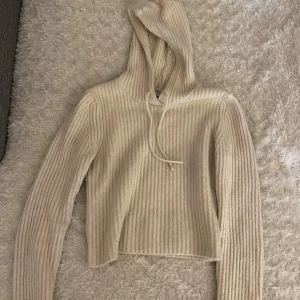 Beige ribbad croppad hoodie - Säljer en beige ribbad hoodie med croppad passform och långa ärmar. Tröjan har huva och är tillverkad i ett mjukt stickat material. 
